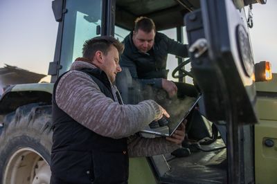 Man with short brown hair,pointing on his laptop while standing next to his colleague,colleague sitting inside his tractor and looking on the screen,in the evening
