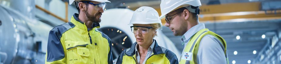 Three Heavy Industry Engineers Stand in Pipe Manufacturing Factory, Use Digital Tablet Computer, Have Discussion. Large Pipe Assembly. Design and Construction of Oil, Gas and Fuels Transport Pipeline