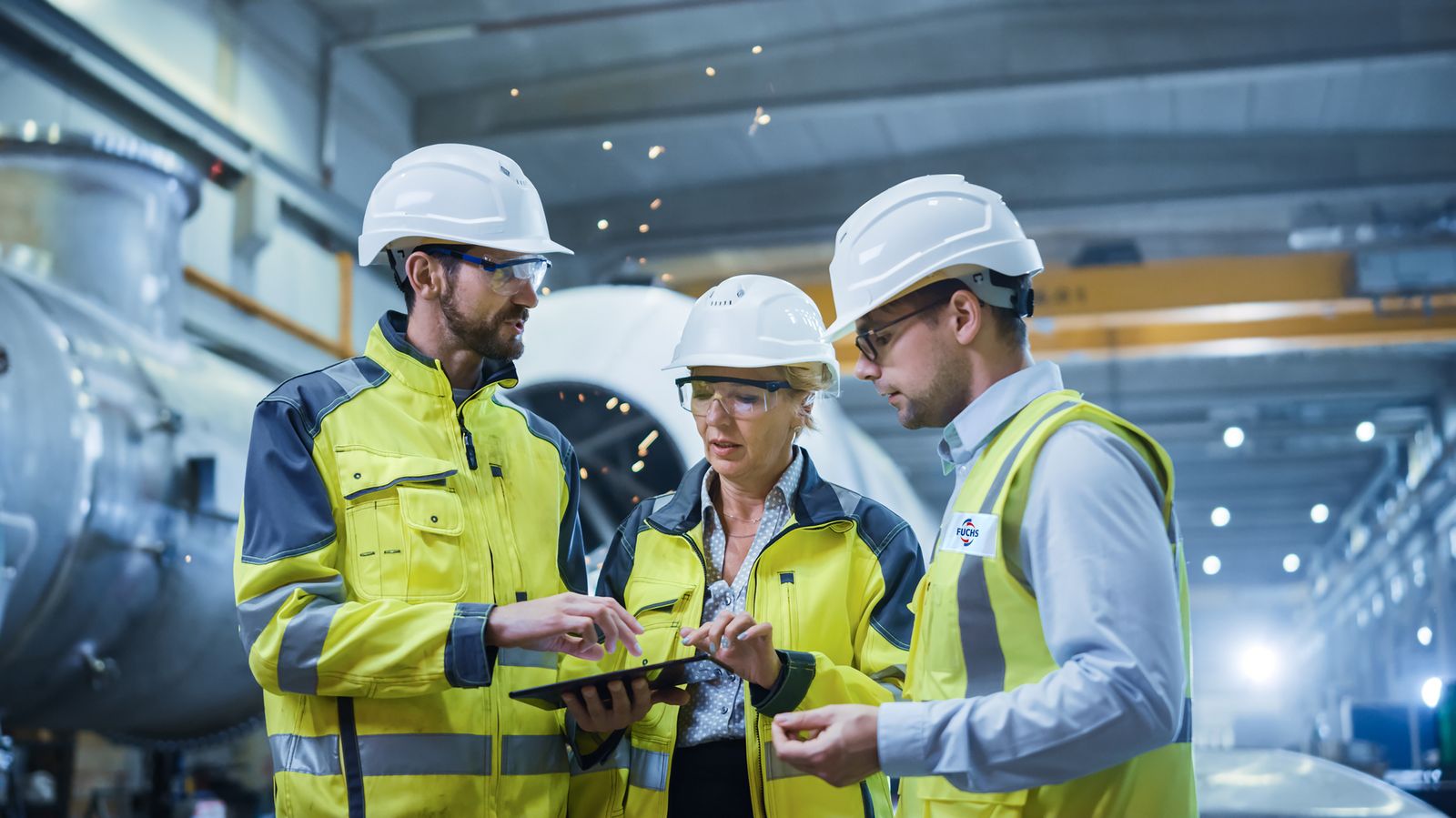 Three Heavy Industry Engineers Stand in Pipe Manufacturing Factory, Use Digital Tablet Computer, Have Discussion. Large Pipe Assembly. Design and Construction of Oil, Gas and Fuels Transport Pipeline