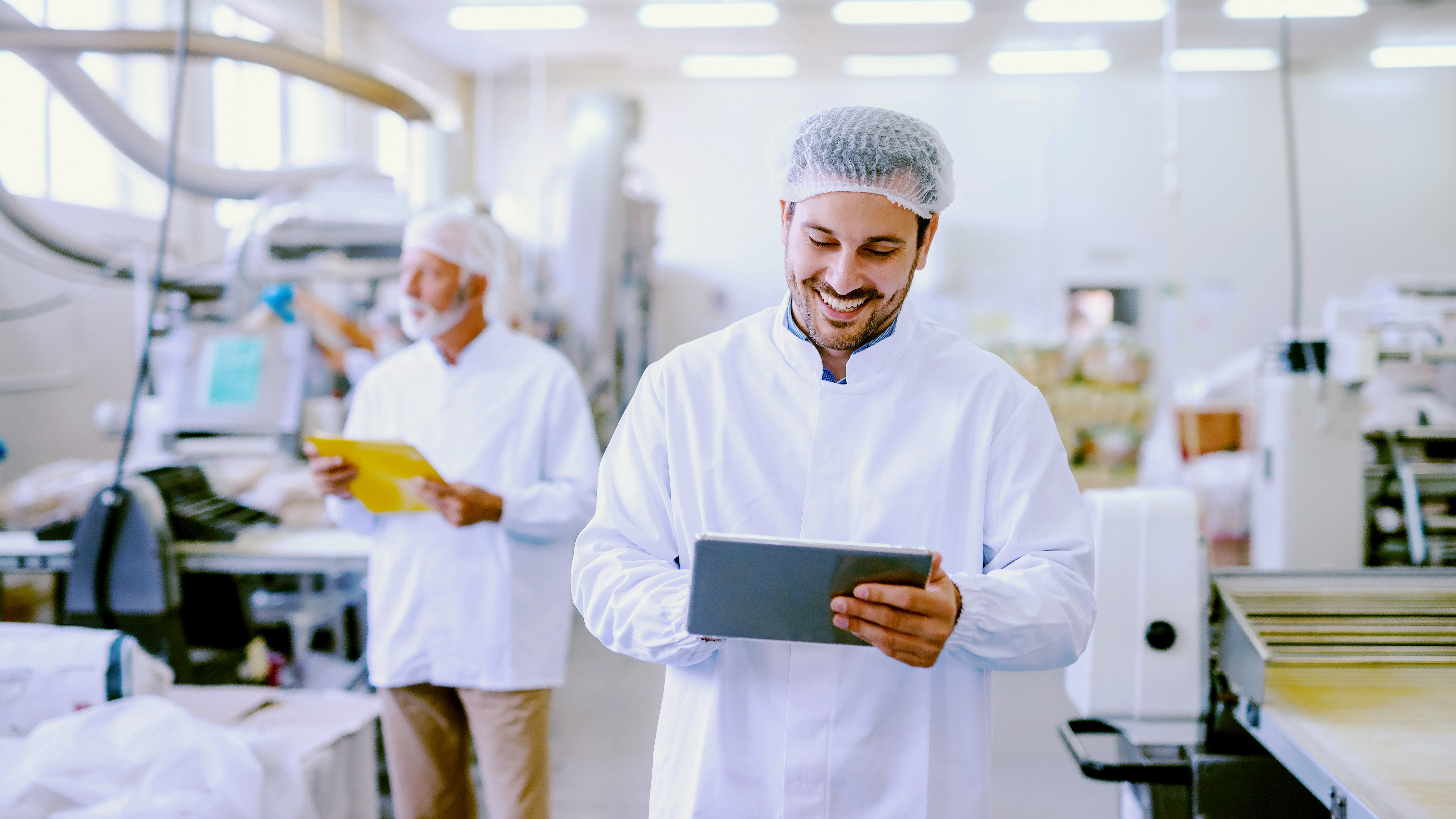 Food production worker wearing protective clothing and a hairnet using a tablet in a modern food manufacturing facility, with another employee and production equipment visible in the background