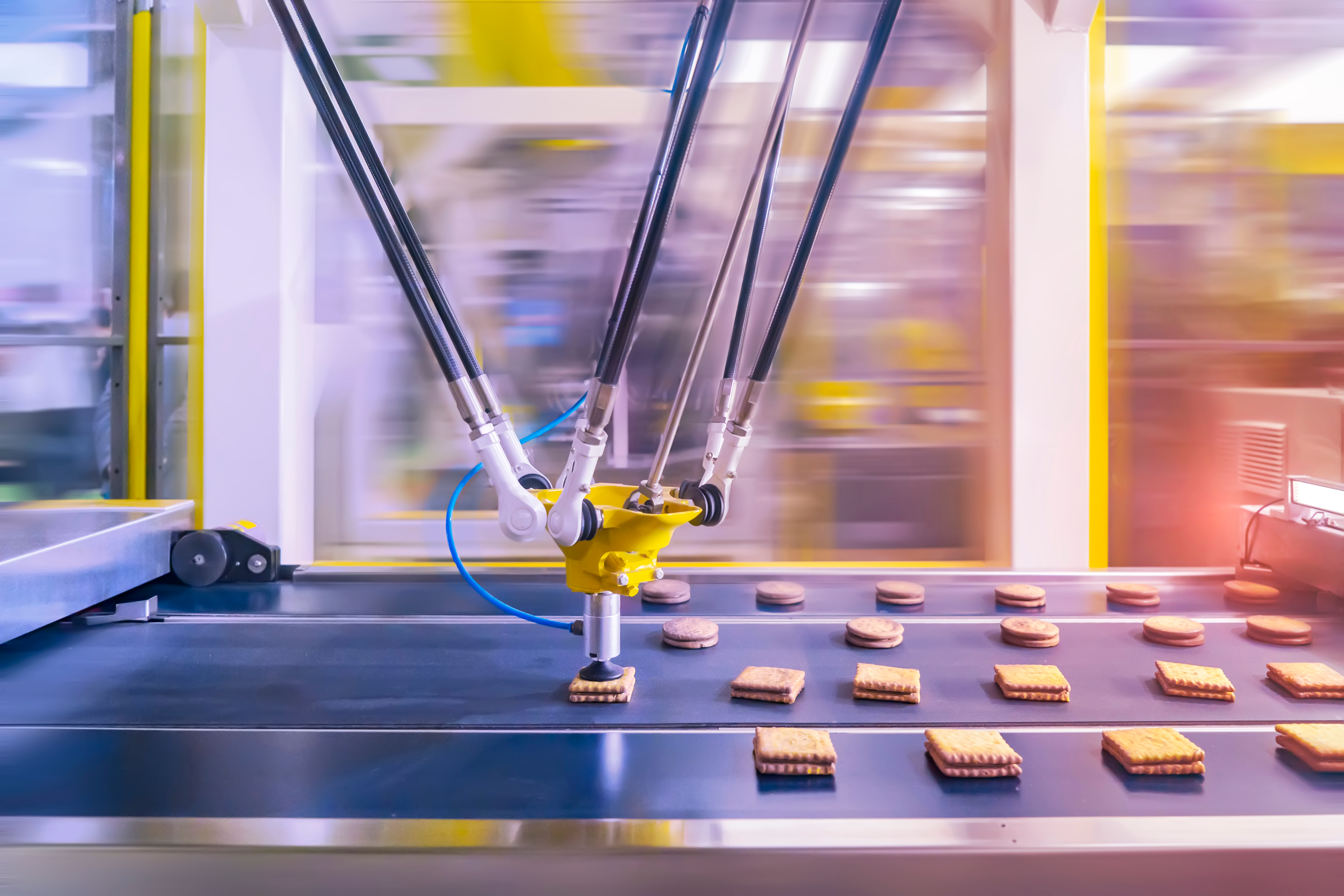 Industrial robotic arm with a white suction cup hovering above chocolate-coated cookies on a conveyor belt in an automated food production line, with additional robotic arms blurred in the background